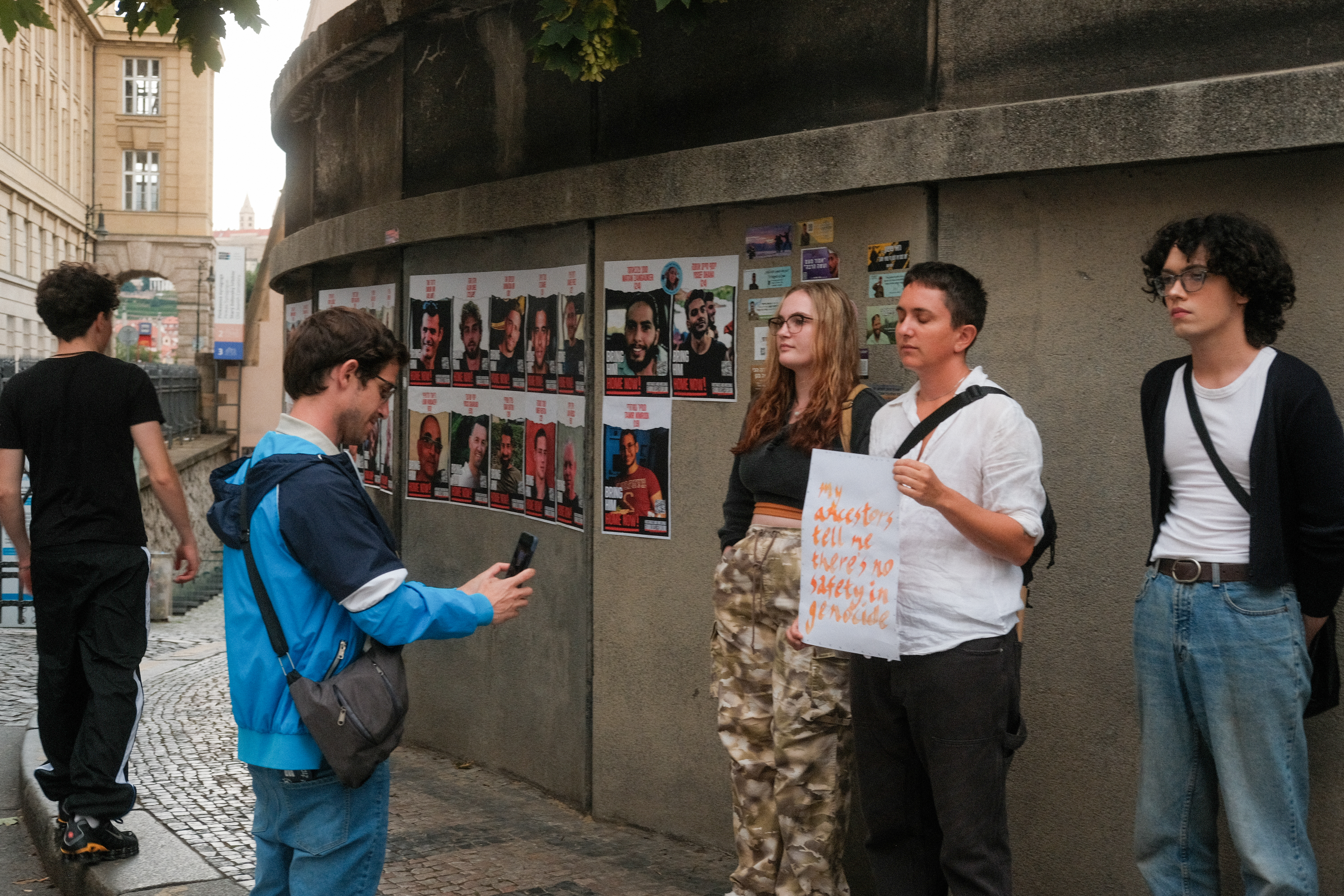 a man photographs three people standing in front of a wall. One holds a sign that reads 'my ancestors tell me there's no safety in genocide.' behind them are pictures of israeli hostages